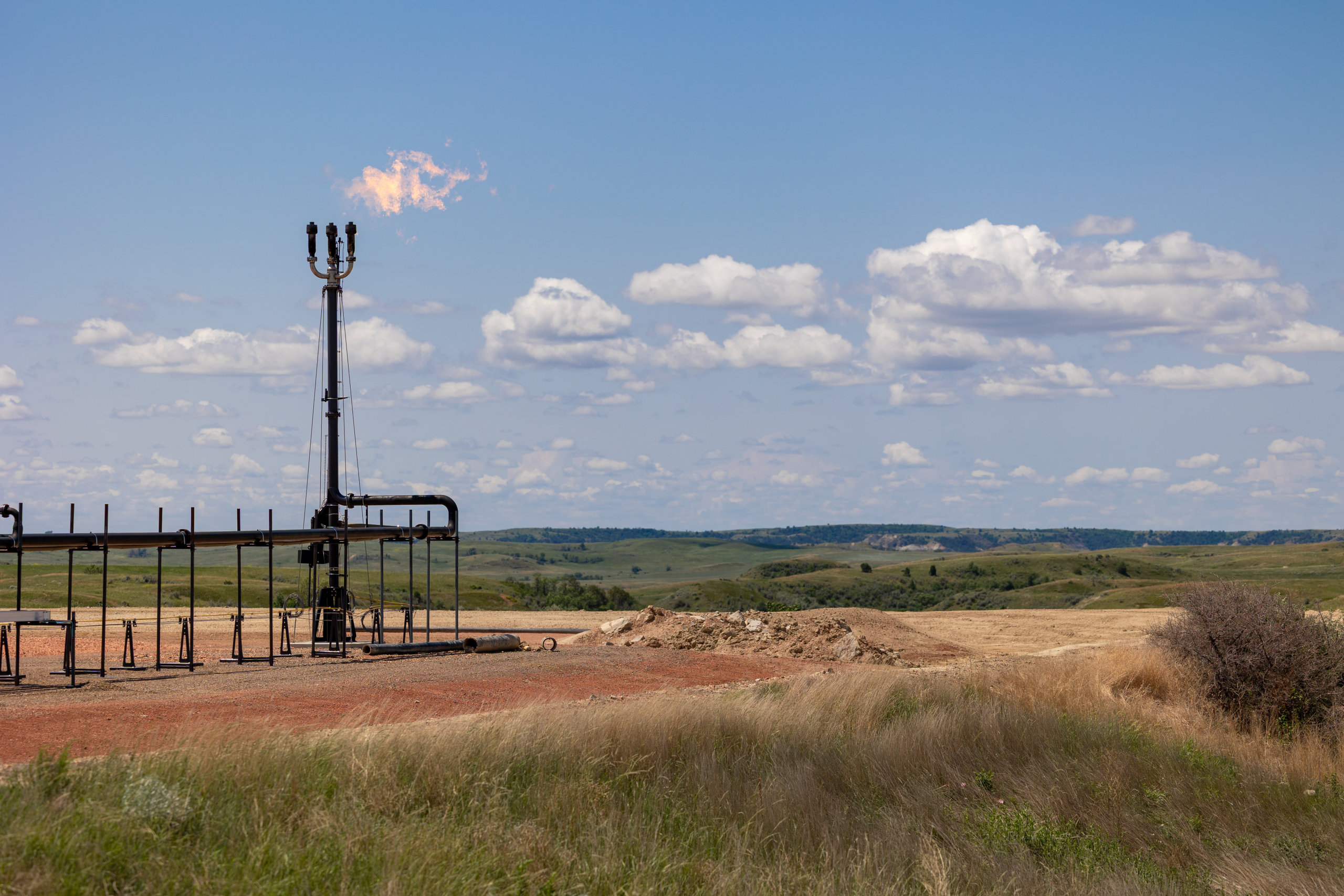 A Variable Orifice Flare burns a clean, smokeless flame under blue skies in the summertime. There is oil and gas equipment surrounding it.